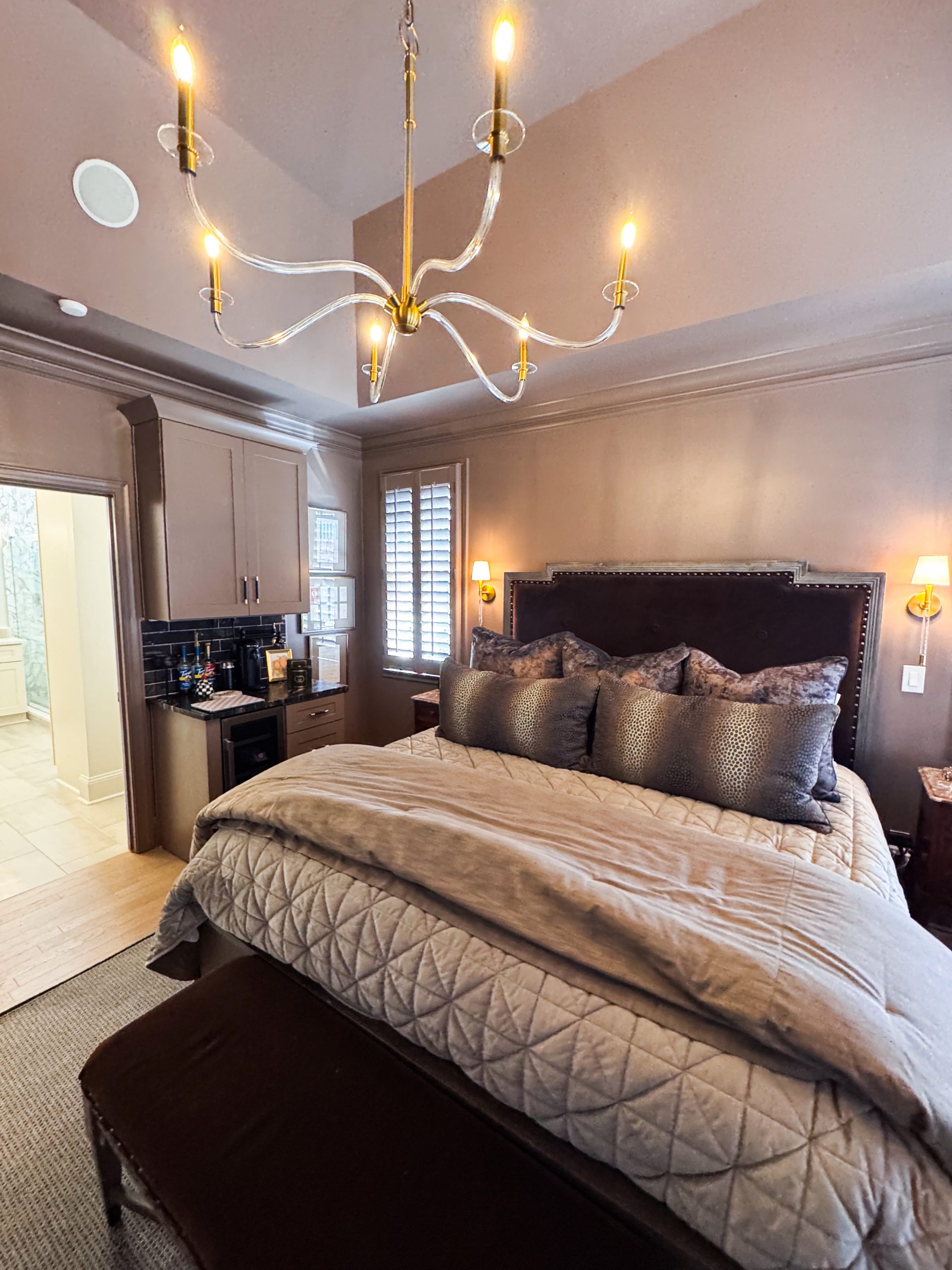 Wide view of the Primary Bedroom with vaulted ceiling, lucite-arm chandelier, upholstered headboard, and antique burl wood chest