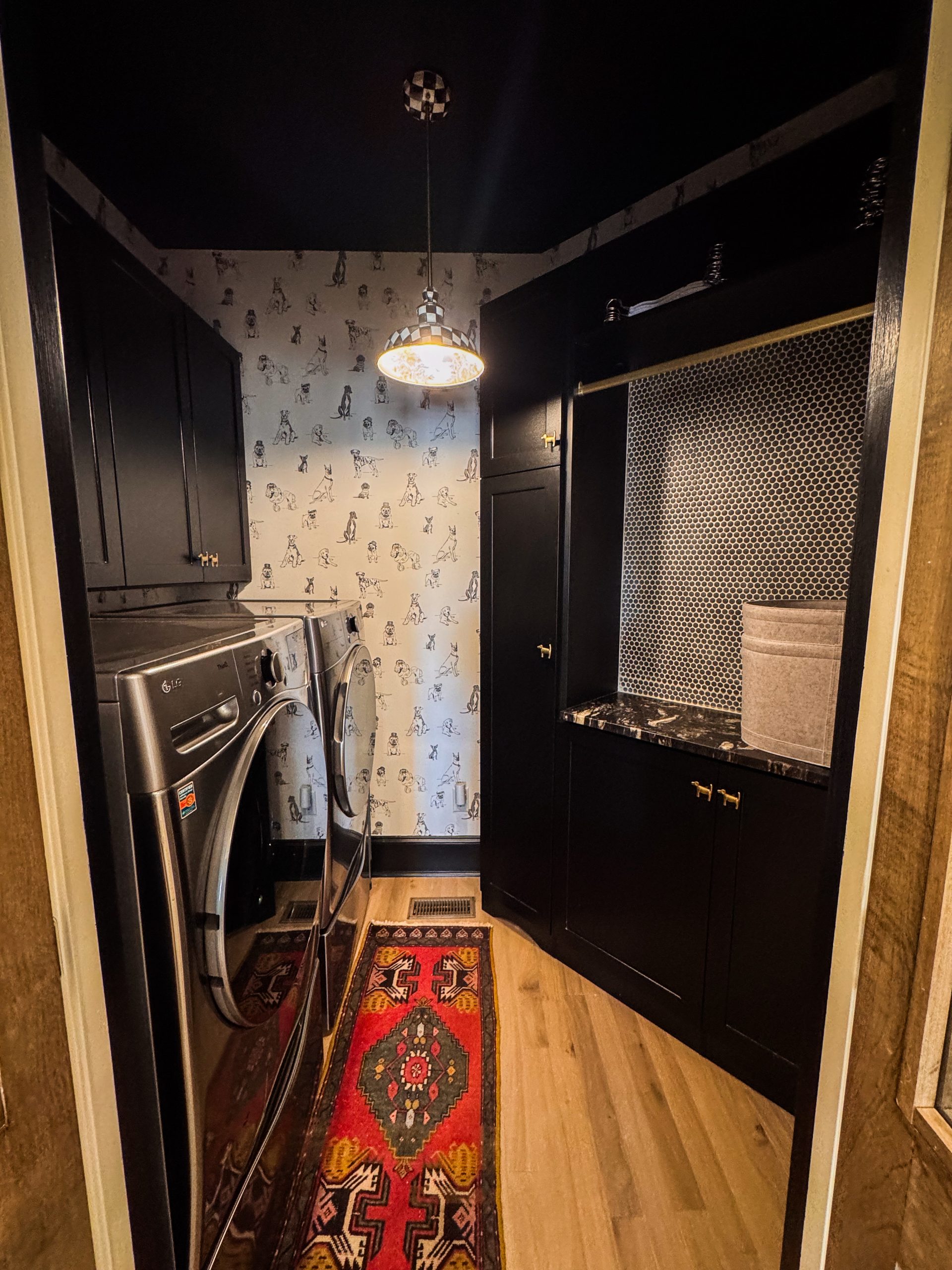 Laundry room wrapped in dog-themed wallpaper with black cabinetry and a hanging rack over a penny-round tile backsplash