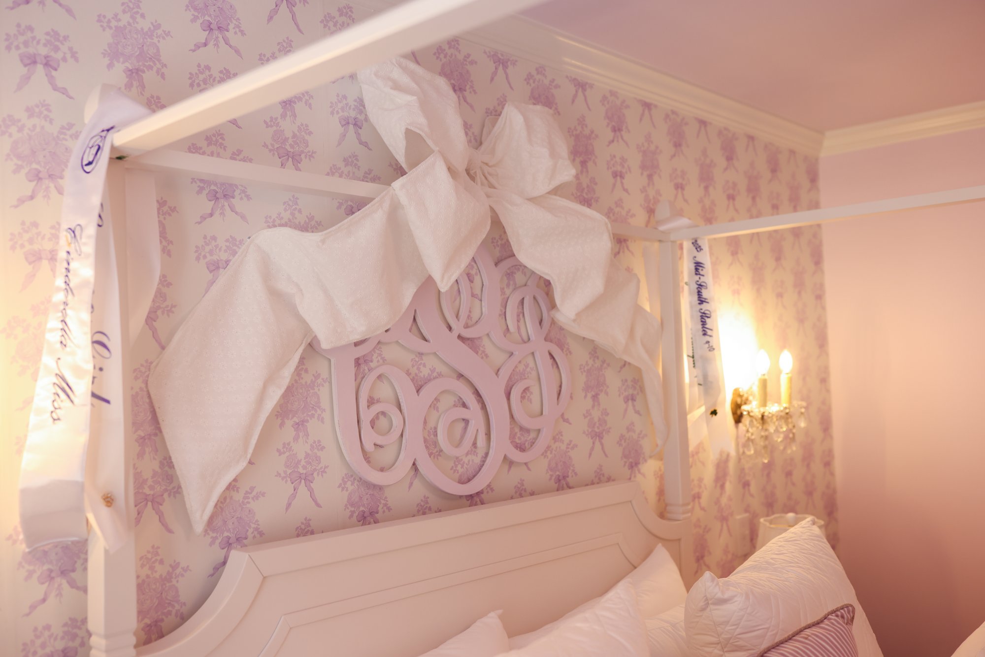 Oversized white bow above the monogrammed headboard beside an antique crystal sconce in Charlotte's bedroom