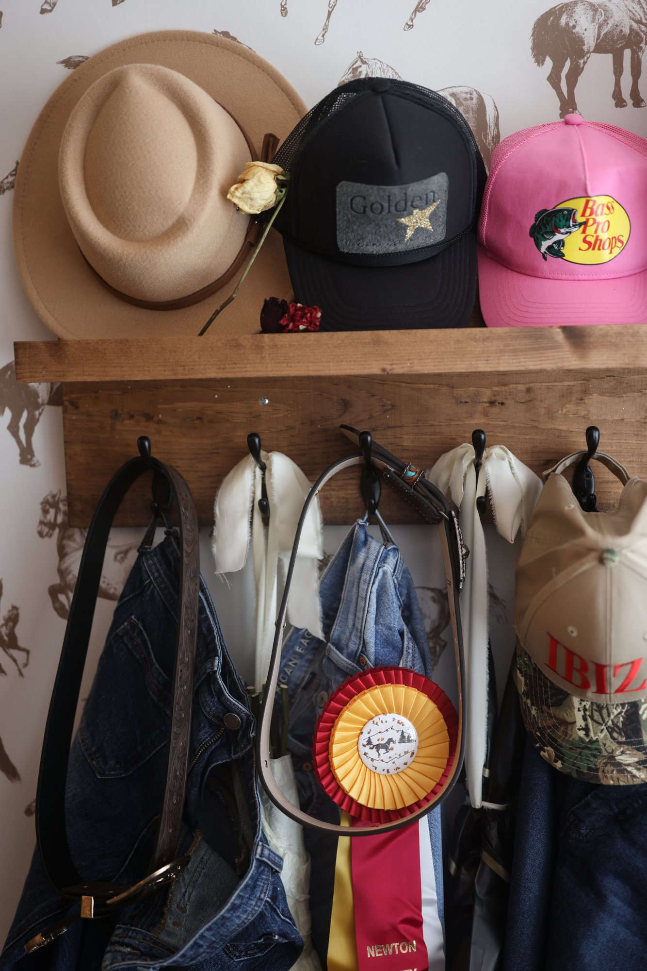 Hat rack with cowboy hats, jeans, and show ribbons against brown quarter horse wallpaper in Caroline's bedroom