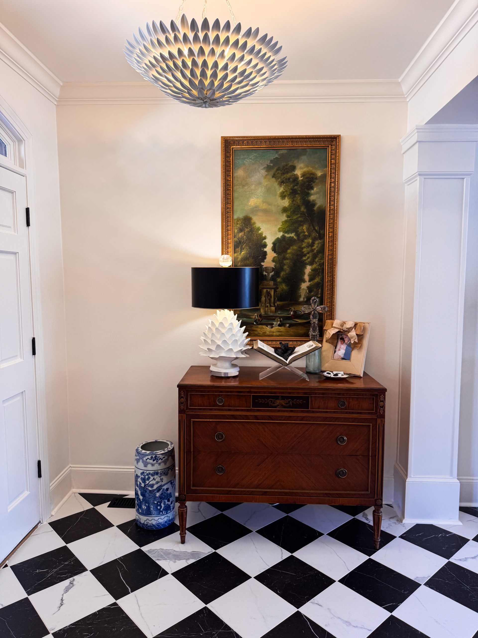 Antique console table beneath a gilt-framed oil painting and sculptural artichoke lamp in a black-and-white tiled foyer