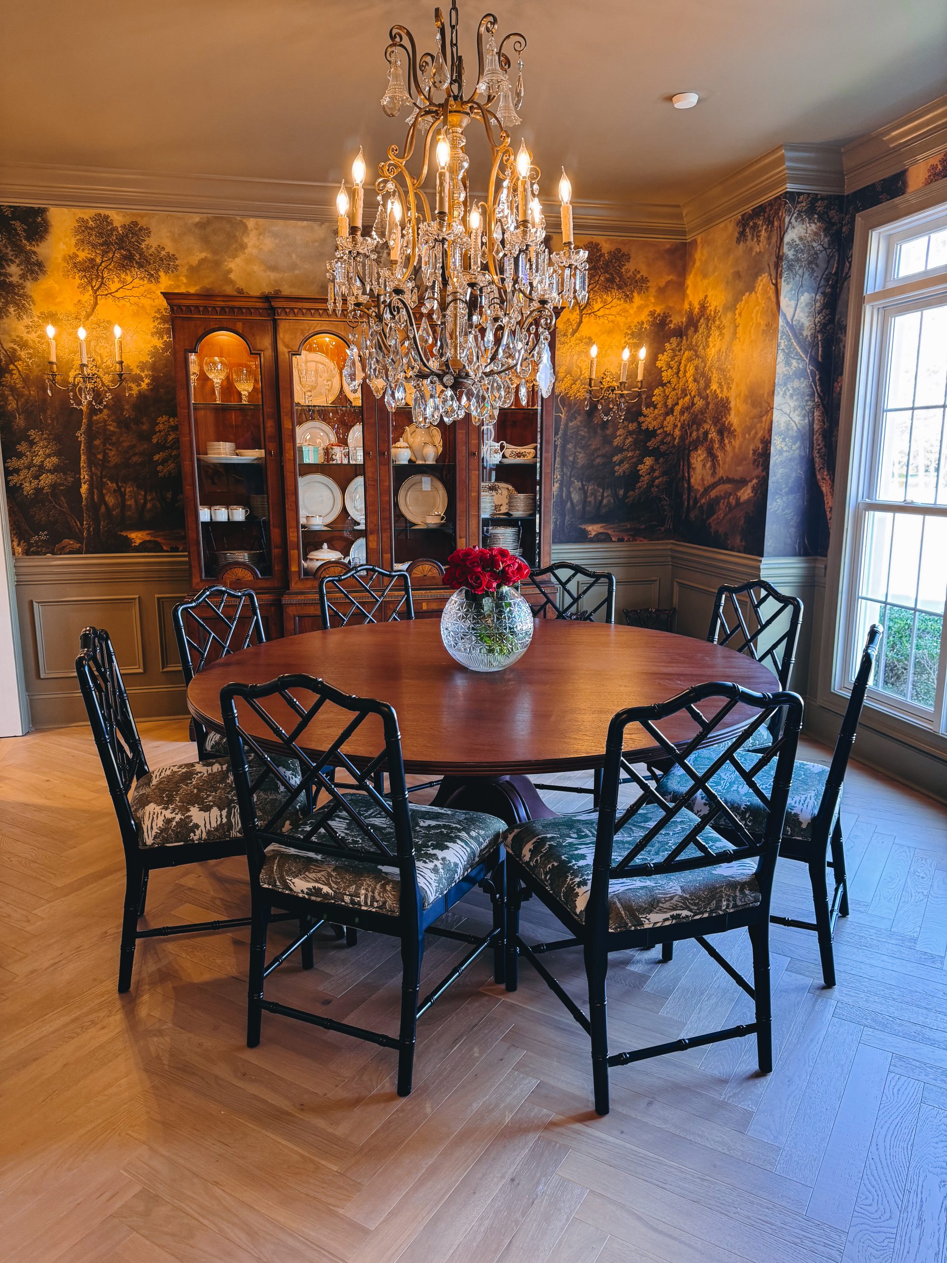 Dining room with a crystal chandelier, round wood table, and scenic mural wallpaper framed by a rich green ceiling and trim